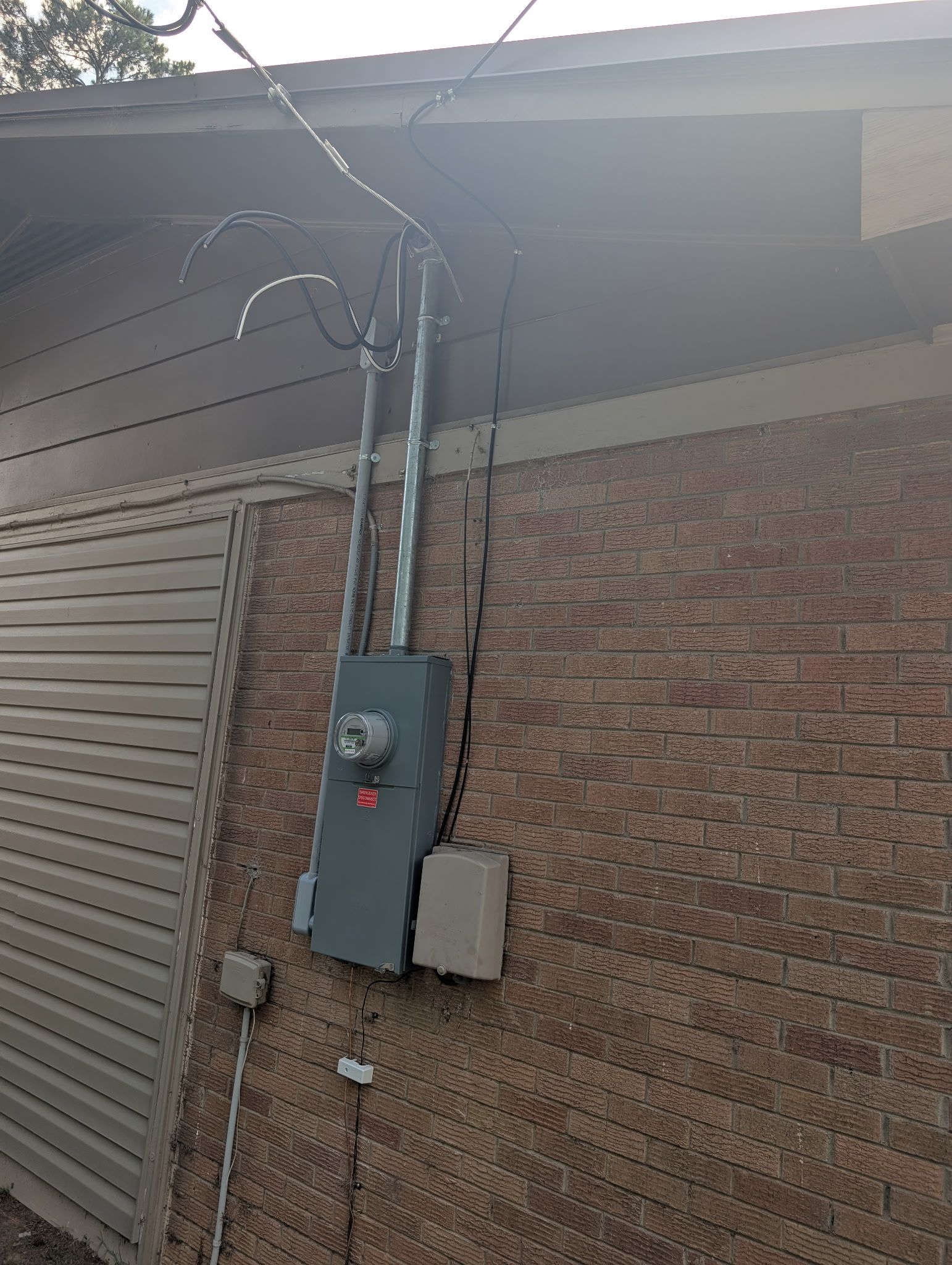 Exterior electrical service on a brick wall — grey meter base and disconnect with a galvanised riser carrying the service conductors up to the roofline